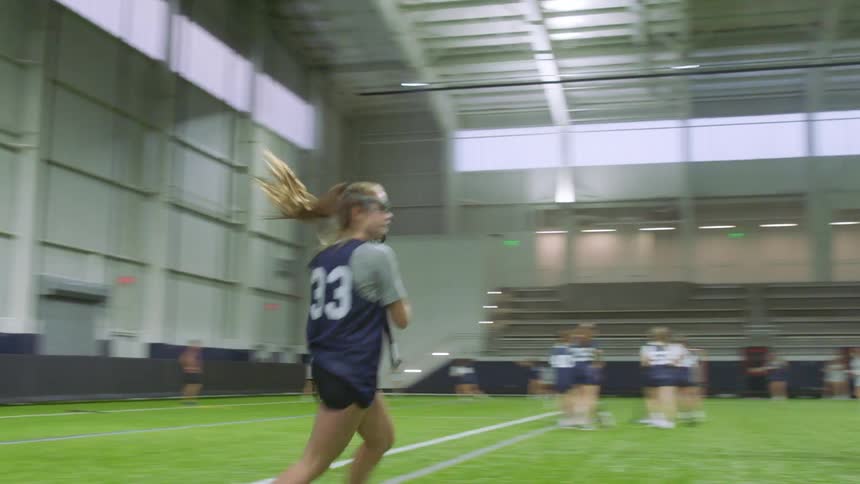 A lacrosse player practices running and catching with her team in an indoor arena