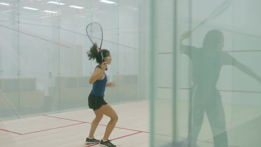 An instructor hits a ball off the wall to a woman in a racket ball court