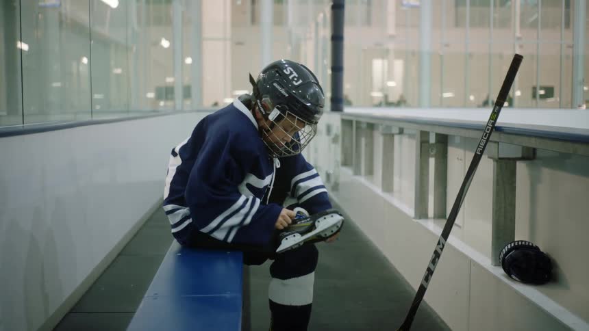 Little boy in hockey uniform tying skates next to ice rink
