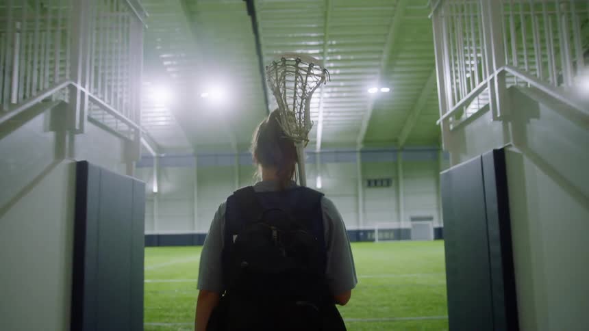 A teenager carrying a lacrosse stick walks out onto an indoor field