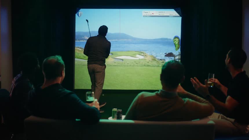 Four men sit on a couch and drink while they watch their friend swing a golf club while playing a video game
