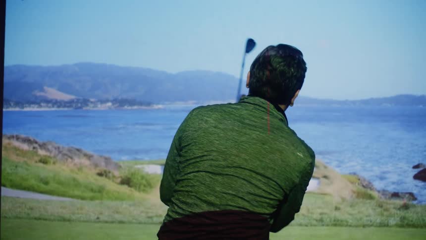 A man plays golf using a large projection screen that shows a coastal golf course