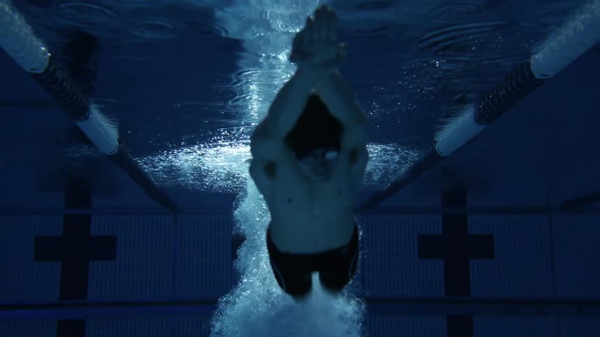 A male swimmer dives into a pool and swims forward underwater