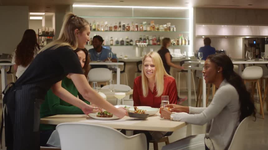 A waitress delivers plates of food to a table of three women happily talking with one another in a restaurant
