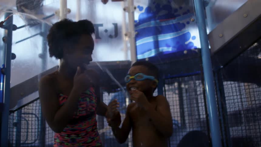 A brother and sister laugh and hug as they stand under spraying water at an indoor water park