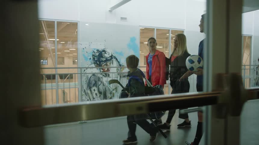 A woman walks through a building with her daughter and sons who carry sports equipment as they walk
