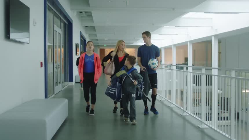 A family walks down the upstairs hallway of a sports complex with one son tossing a soccer ball in the air and another son holding a hockey stick