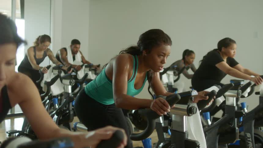 A young black woman determinedly pedals as she rides a stationary exercise bike beside others in a fitness center