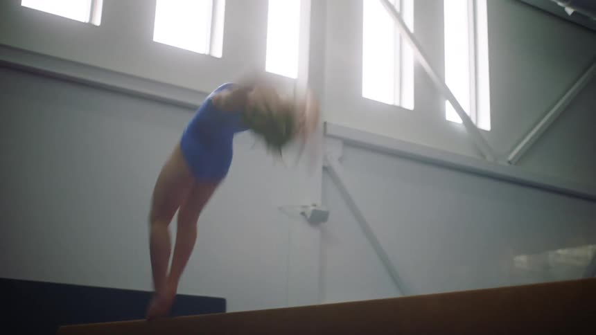 A female gymnast standing on a balance beam attempts a backflip but fails and falls off