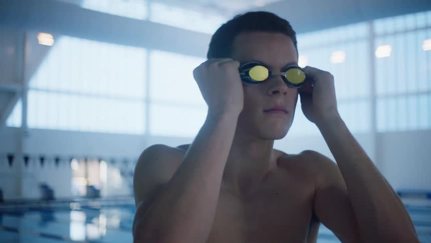 A man standing and putting on goggles with a pool and windows in the background