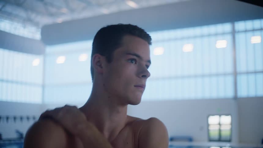 A shirtless young man does stretches while standing on the side of an indoor swimming pool