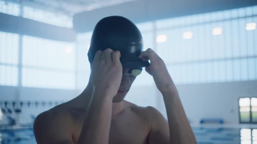 A swimmer puts on a swim cap while wearing goggles and standing by the indoor pool