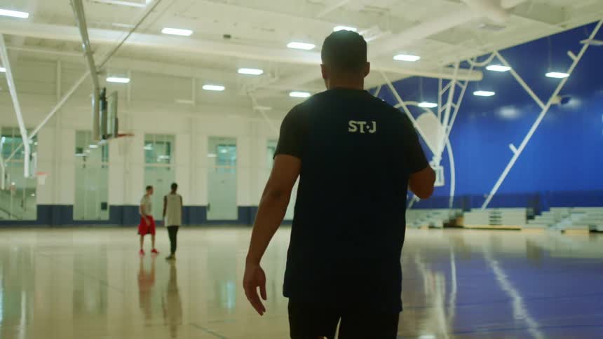 A young man dribbles a basketball as he walks across a gymnasium towards a group of young men practicing their shooting