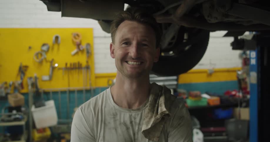 Mechanic working in a workshop under a car puts a towel on his shoulder and looks smiling at the camera
