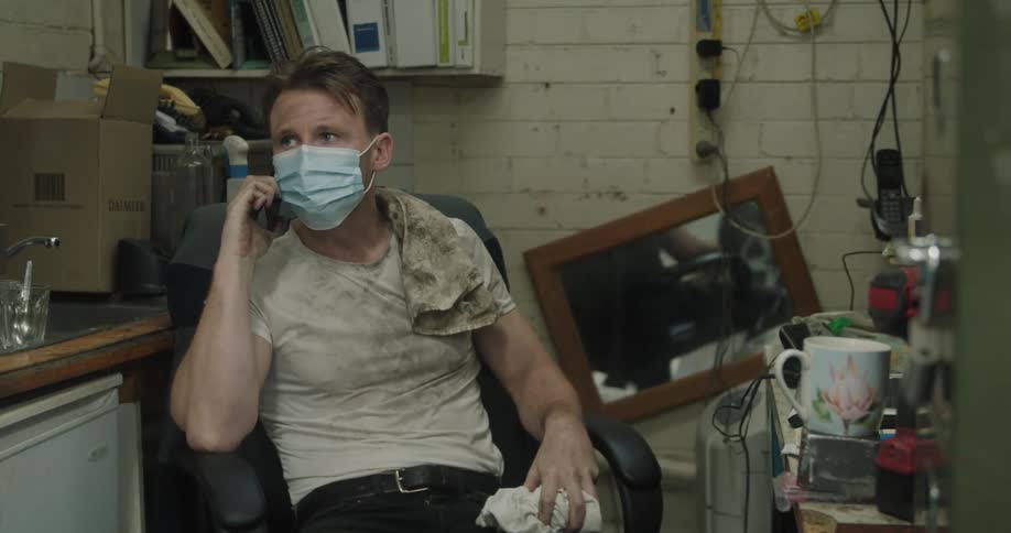 Car mechanic with a blue mask and a dirty white t shirt sits in an armchair and talks on smartphone in a room