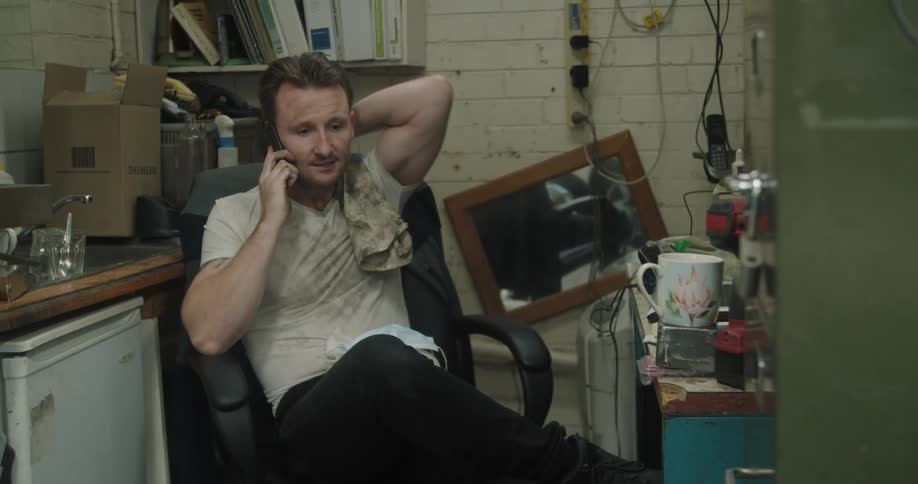 A man in a dirty shirt talks on a smartphone while sitting in a chair in a small junky room