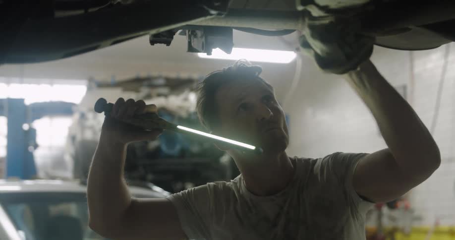 A mechanic stands under a raised car and examines it with a light