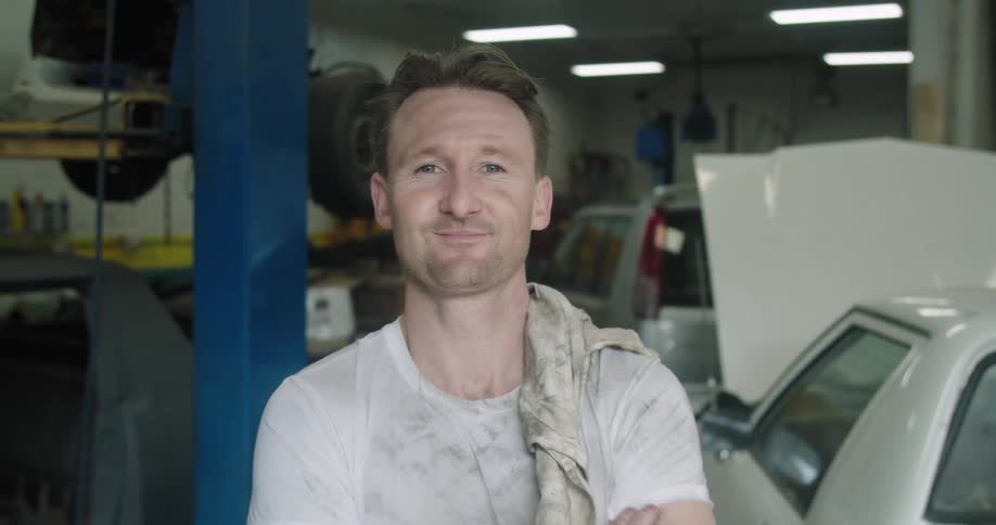 Car mechanic with a dirty white t shirt throws a dirty rag over his shoulder and looks at the camera smilingly in the garage