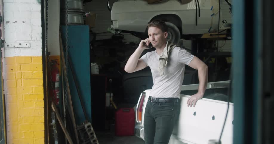 A mechanic leans against a car talking on his cell phone just inside a garage