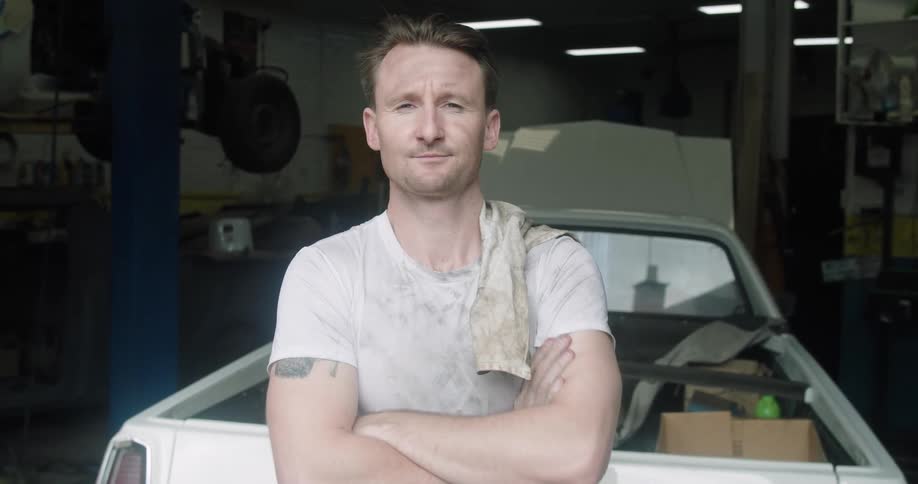 A mechanic stands just outside of a garage and flips a towel over his shoulder