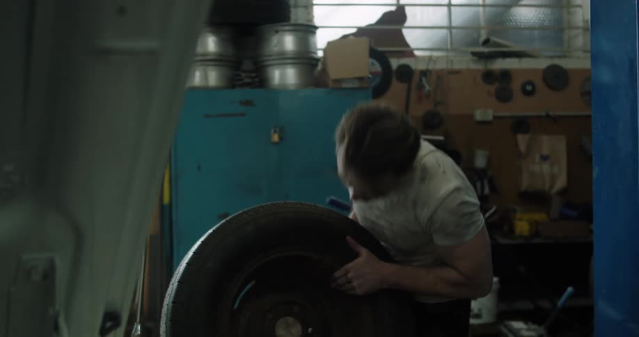 Car mechanic with a dirty t shirt lifts a tire up in the garage