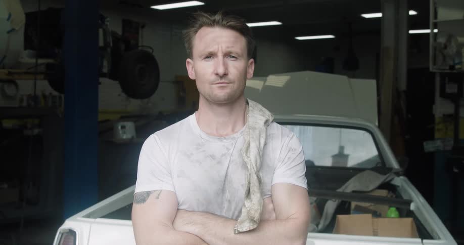Car mechanic with a white dirty t shirt stands arms crossed and faces the camera with eye contact in the garage