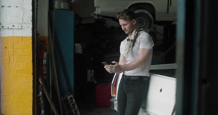 Car mechanic with a dirt t shirt and jeans uses a smartphone in the garage