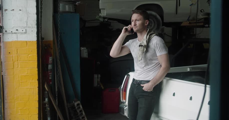 Man leans against a truck in a vehicle repair shop and talks on a cell phone