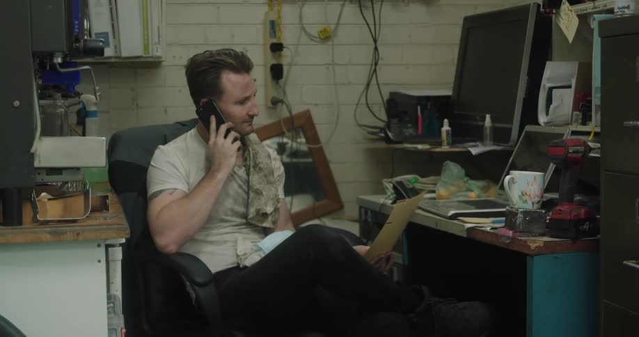 Car mechanic with a dirty white t shirt sits in an armchair and talks on smartphone in a room