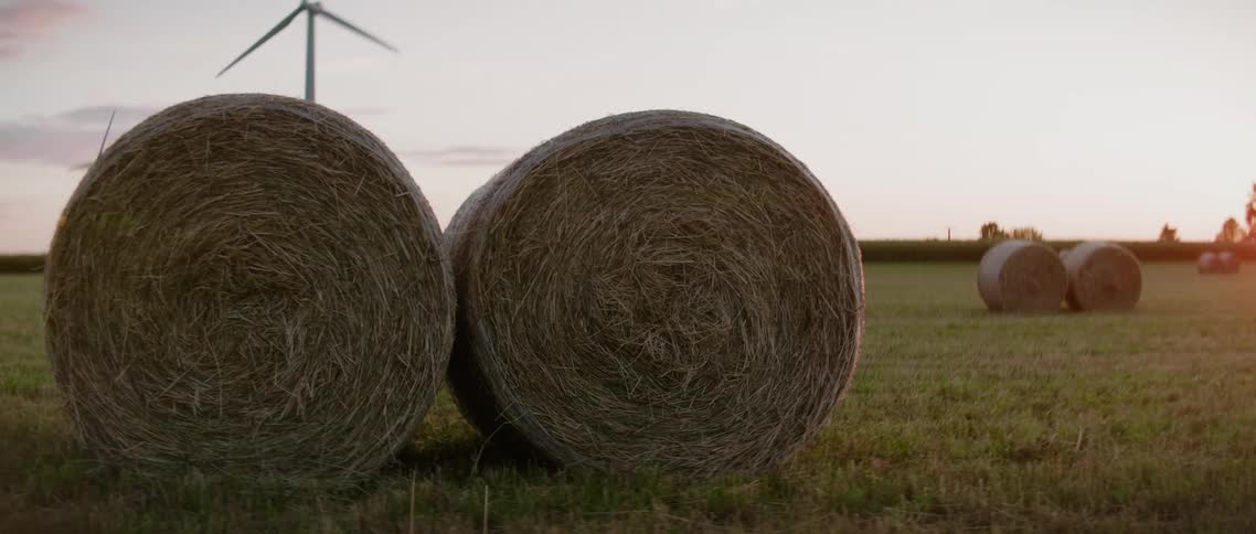 Pairs of hay bales lie in the field of a wind farm during the day
