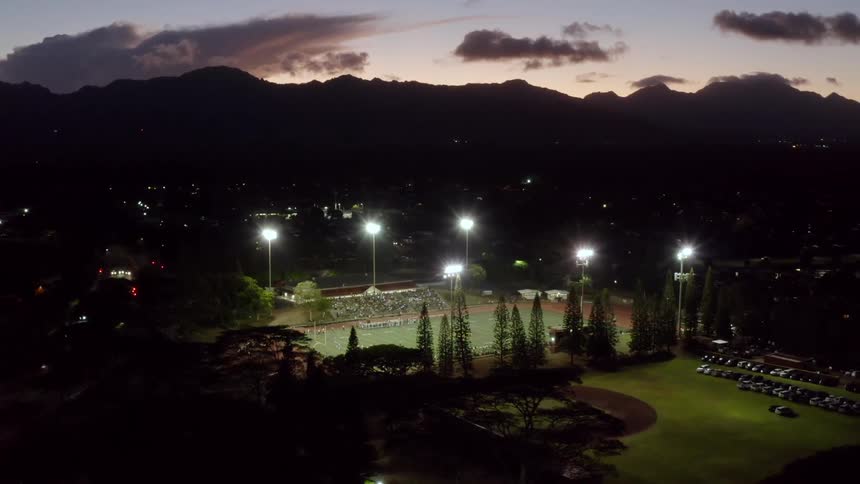 A football stadium is illuminated by flood lights as spectators watch the football players gathered on the field at night