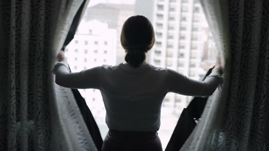 A woman opens the drapes in her apartment and looks out the window