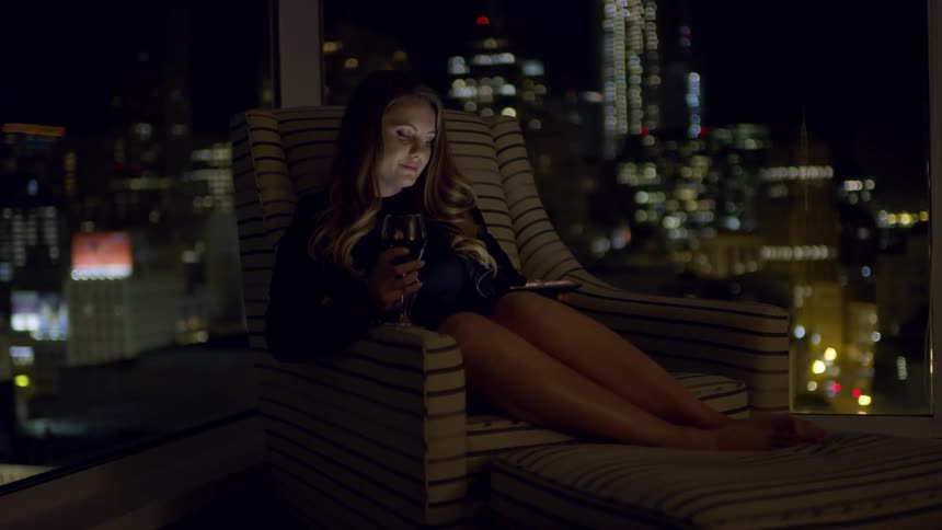A woman sits in a striped chair in her high rise condo downtown and uses her smartphone while holding a glass of red wine