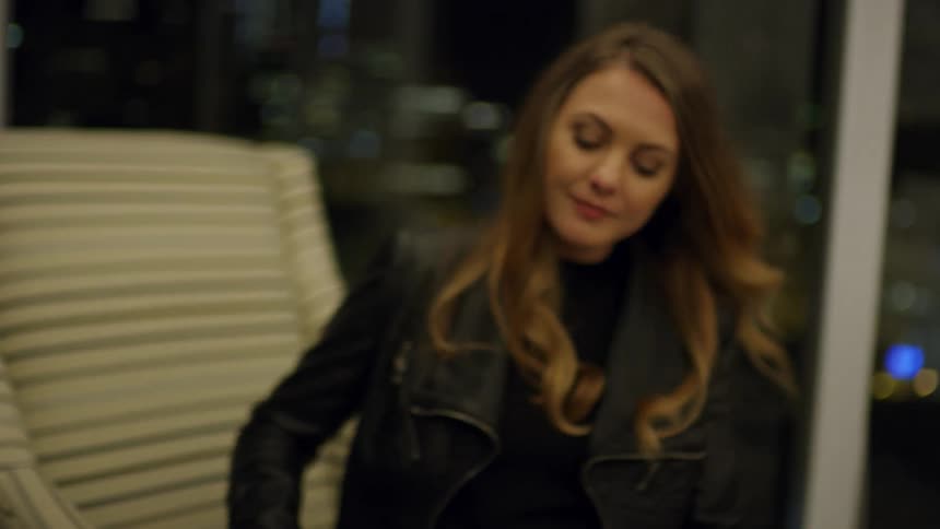A woman in her home walks to a chair and sits down and takes her shoes off
