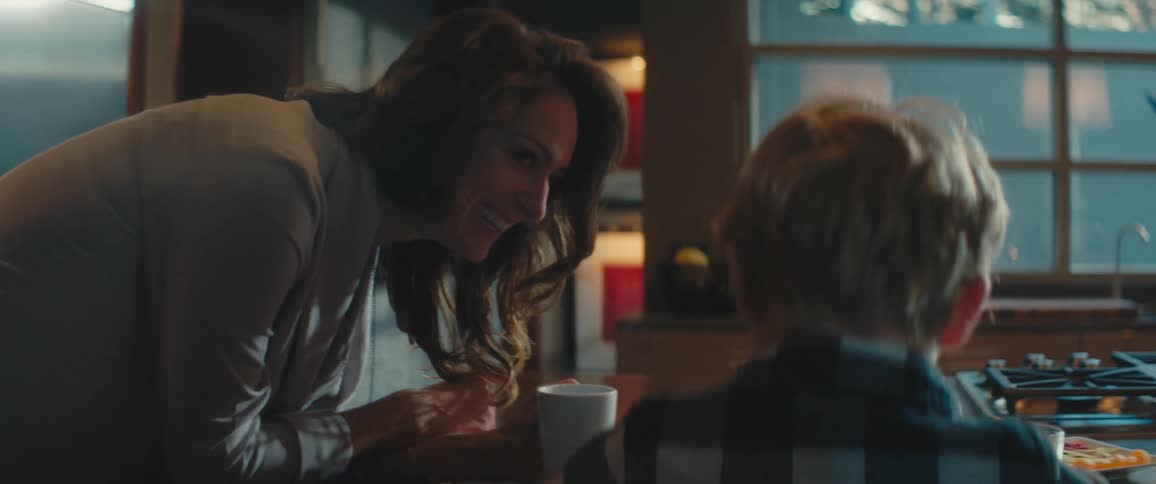 A young boy sits at a kitchen island with his mother leaning beside him and laughing