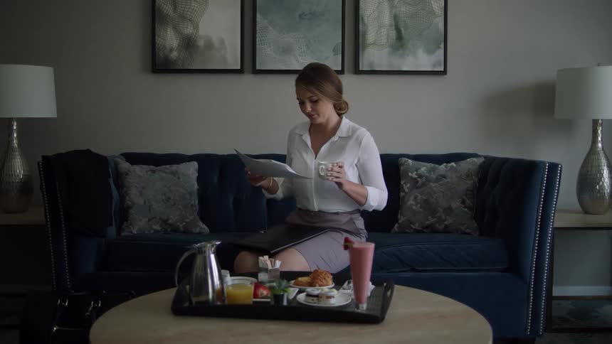 A woman sits on a couch and reads some papers while picking up a beverage from a tray with food and juice on it