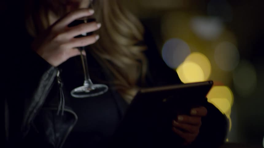 A woman drinks a glass of wine while looking at her tablet