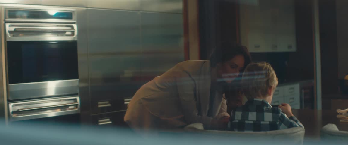 A boy sits in a kitchen with an electronic device and his mother leans over him