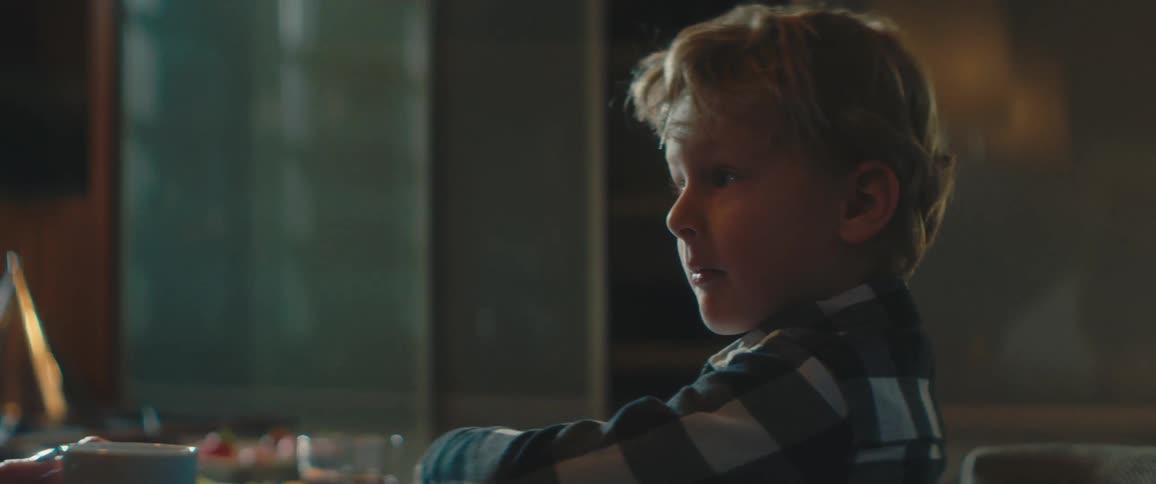 A young boy sitting at a dining table looks up from a tablet at a person sitting just out of frame