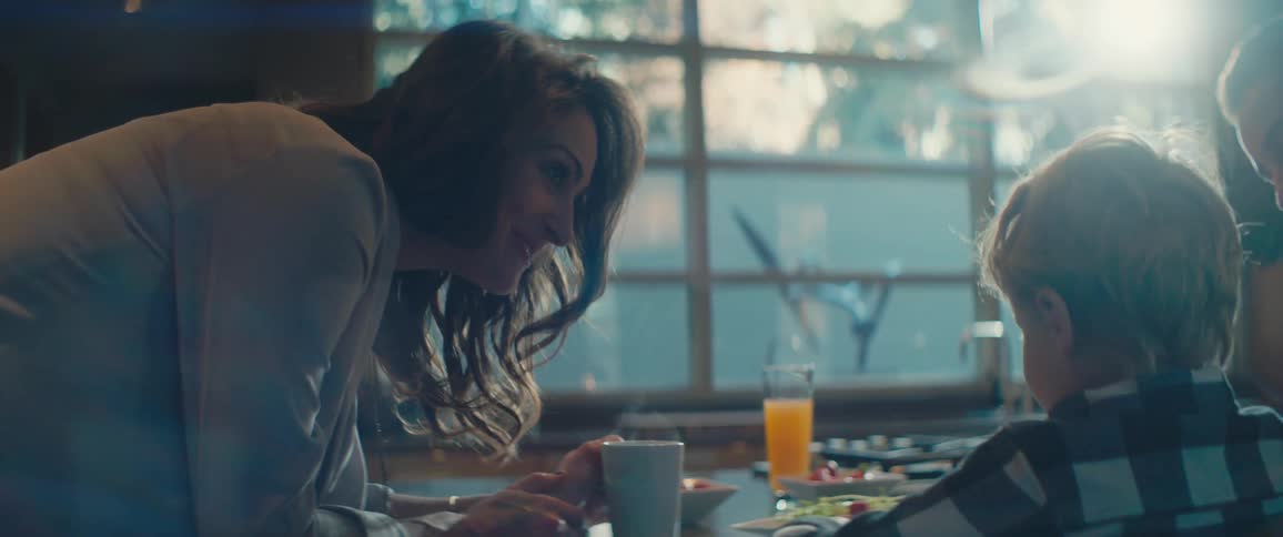 A woman leans down on a table and talks and laughs with her son who is eating breakfast with his dad