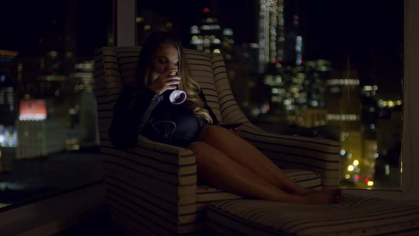 Woman with brown hair sits in armchair drinks a glass of wine while using smartphone in a room with city skyline in the background at night