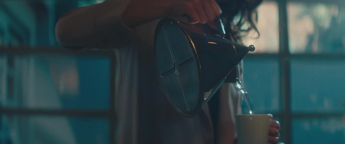 A woman pours steaming water from a kettle into a coffee cup and smiles as she walks out of frame