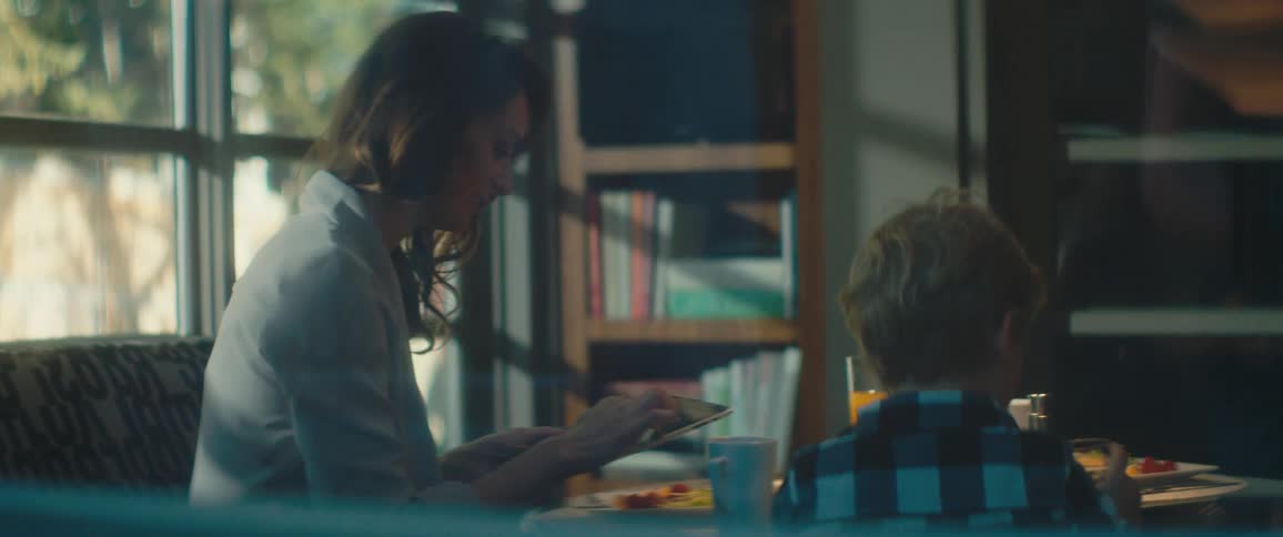 A woman uses a tablet while sitting at a dining table with a boy who drinks from a glasses
