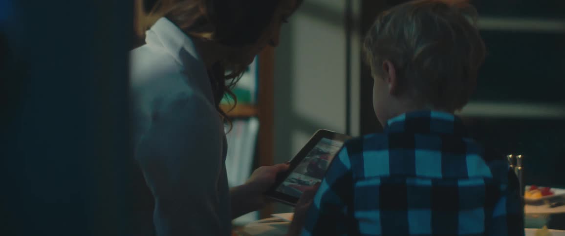 A boy points at the screen of a tablet being held by his mother as they sit together at a dining table