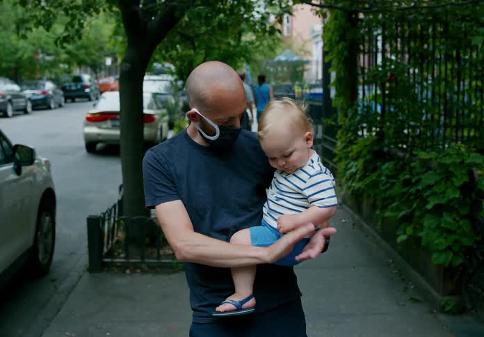 A man in a medical mask claps while holding a baby with one arm as he stands on the sidewalk in a city neighborhood