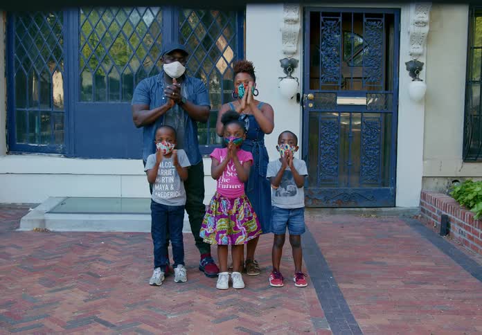 Family wearing colorful face masks standing outside their home clap while looking at the camera