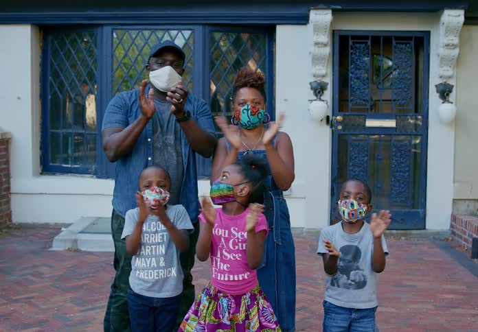 A black family wearing medical masks claps while standing outside of house and looking into the camera