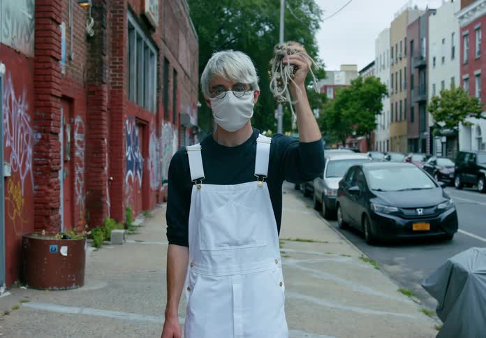 A young man in white overalls shakes a string of bells while standing on a city sidewalk during the day