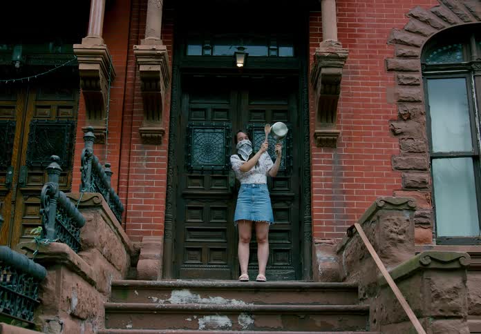 A woman stands on a front porch using a wooden spoon to bang a metal pot