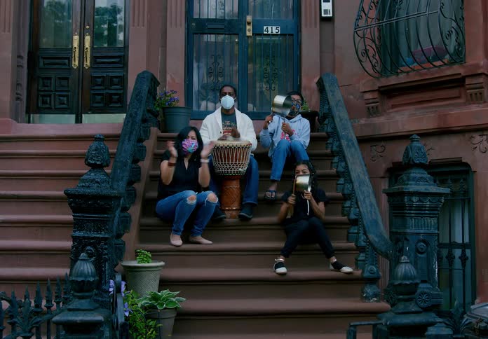 A family claps and bangs pots and drums while sitting on the stairs outside of a building